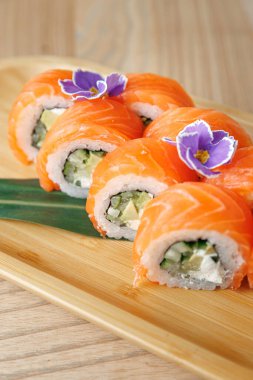 Suşi ruloları ahşap bir tabakta güzelce düzenlenir. Her rulo somon, salatalık ve avokado içerir. Yenilebilir çiçekler sunuma renk ekler.