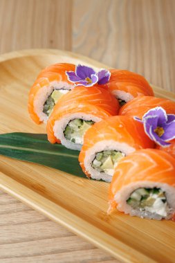 Salatalık ve krem peynirle dolu suşi ruloları zarif bir şekilde ahşap tabağa konur. Yenilebilir çiçekler dekoratif bir dokunuş ekleyerek bu Japon yemeğinin sunumunu güçlendirir..