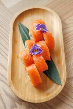 Pirinç ve sebzeyle doldurulmuş suşi ruloları taze somonla kaplı ahşap bir tepsiye yerleştirilir. Yenilebilir çiçekler ve yeşil yapraklar yemeğe biraz renk katar..