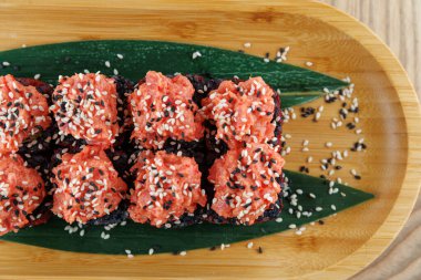 Suşi börekleri, üzerine baharatlı somon karışımı ve susam tohumları serpiştirilmiş ahşap bir tabakta oturur. Tabakta garnitür için yeşil yapraklar var. Renkli bir yemek sunuyor..