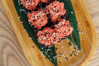 Suşi rulolarında pembe deniz ürünleri ve siyah pirinç üzerine susam tohumları bulunur. Bir restoranın ahşap tepsisine yerleştirilmiş yeşil bir yaprağa yerleştirilmişler..