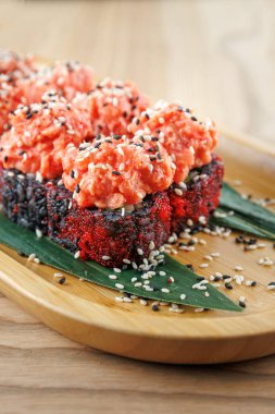 Suşi ruloları ahşap bir tabakta güzelce düzenlenir. Ekmekler somon ve susam tohumlarıyla kaplı. Dekoratif yeşil yapraklar sunuma biraz renk katar.