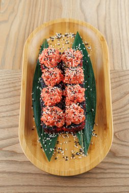 Suşi ruloları üzerine susam tohumları serpiştirilmiş bambu bir tabakta düzenlenir. Çanak, ahşap masa yüzeyinde renkli bir sunum sergiliyor..