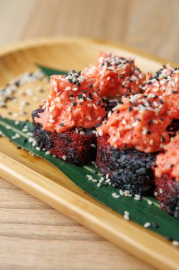 Suşi ruloları ahşap bir tabakta yeşil bir yaprağa yerleştirilir, üzerine baharatlı somon karışımı ve susam tohumları serpiştirilir. Basit ve çekici bir yemek sunumu gösterilir.