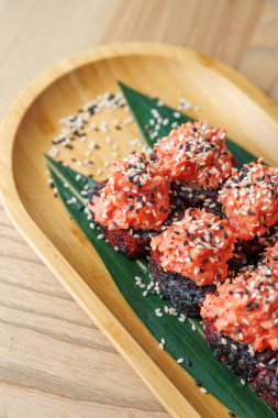 Üzerine pembe somon ve susam tohumları serpiştirilmiş suşi ruloları ahşap bir tepside bambu yaprağının üzerinde duruyor. Bu yemek lezzet ve dokuların bir karışımını sergiler..