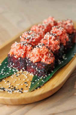 Pembe deniz ürünleri soslu taze suşi ruloları ahşap bir tabakta sergilenir. Ekmekler susam tohumlarıyla süslenip muz yaprağına konuyor. Paylaşım için mükemmel..