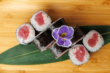 Pilav ve ton balığıyla yapılan taze suşi ruloları bambu tahtasında güzelce düzenlenir. Dekoratif bir mor çiçek en üste yerleştirilir, sunuma biraz renk katar.