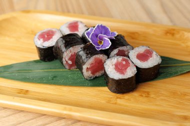 Suşi ruloları bambu bir tabakta, içinde pirinç ve deniz yosunlarına sarılmış balıklar var. Mor bir çiçek, ahşap bir masanın sunumuna bir dokunuş ekler..