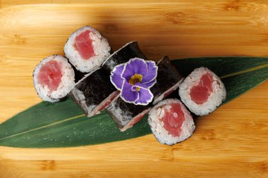 Bir bambu tabağında ton balığıyla dolu beş suşi rulosu düzenlenir. Mor bir çiçek, Japon mutfağında kullanılan taze malzemelerin altını çizerek yemeğe renk katar..