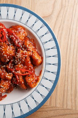 Kırmızı biber ve susam tohumlarıyla süslenmiş tatlı ve baharatlı bir sosla kaplı bir tabak karides tutar. Tabak canlı renklere sahiptir ve açık ahşap bir masaya dayanır..