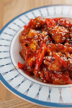 Bu sahnede mavi ve beyaz tabakta servis edilen tatlı ve baharatlı tavuklar yer alıyor. Canlı yemek susam tohumları ve kırmızı biber dilimlerini içeriyor. Ortam davetkar ve sıcak, yemek için mükemmel..