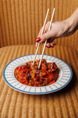 A hand holds chopsticks while picking up colorful stir-fried vegetables from a round plate. The setting features a woven texture, adding warmth to the scene.