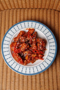 Susam tohumu ve kırmızı biberle süslenmiş baharatlı sosta pişirilmiş bir gurme tavuk tabağı dekoratif bir tabakta sunulur. Ayarda hasır kaplamalı bir arkaplan var.