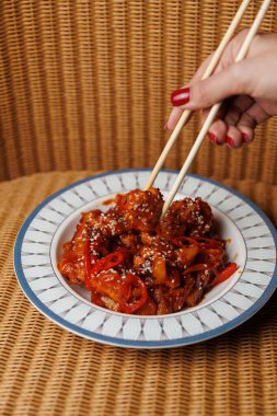 A hand is lifting a piece of meat with chopsticks from a bowl filled with cooked meat, vegetables, and sauce on a textured brown background.