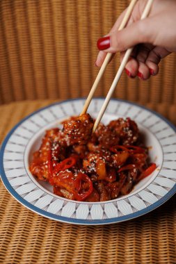 A hand holds chopsticks to lift a serving of meat and vegetables coated in a rich sauce. The dish is garnished with sesame seeds and presented on a decorative plate against a woven background.