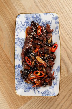 Sığır eti sebze ve acı biber dilimleriyle kızartılır ve susam tohumlarıyla kaplanır. Tabak, ahşap bir yüzey üzerinde duran dekoratif bir tabakta servis edilir..