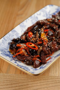 Bir tabakta et, sebze ve baharat karışımı bulunur, dilimlenmiş kırmızı biber ve susam tohumlarıyla süslenir. Tabakta çeşitli elementleri desenli bir tabakta birleştiren zengin renkler ve dokular yer alıyor..