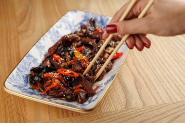 Chopsticks grasp a serving of stir-fried meat with colorful vegetables on a decorative plate. The setting is a wooden dining table.