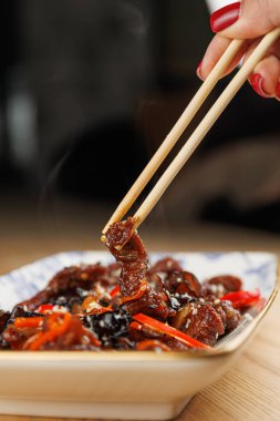 A hand lifts a piece of tender beef stir-fry with colorful vegetables using chopsticks from a plate. The dish emits steam on a wooden table during a mealtime.