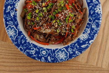 Bir kase kızarmış şehriye renkli sebzeler ve dekoratif bir tabakta sıralanmış susam tohumları içerir. Bu yemek Asya mutfağından bir tat sunuyor..