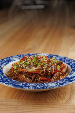Renkli sebzelerle karıştırılmış soba erişteleri dekoratif mavi ve beyaz bir kasede otururlar. Tabaktan buhar yükseliyor, sıcak bir yemek alanında sıcak bir atmosfer yaratıyor..