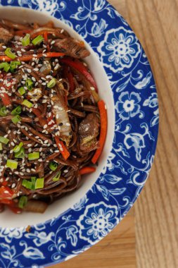 Bu tabak erişteleri sebzelerle karıştırıp susam tohumları ve yeşil soğanlarla süslüyor. Renkli sunum dekoratif bir kasede lezzet ve dokuların bir karışımını vurguluyor.