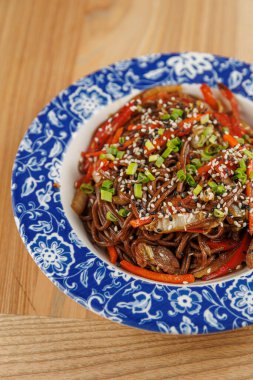 Soba şehriyeleri kırmızı biber ve susam tohumları ve doğranmış yeşil soğan gibi renkli sebzelerle karıştırılır. Bu tabak ahşap bir masada mavi bir kaseye konur..