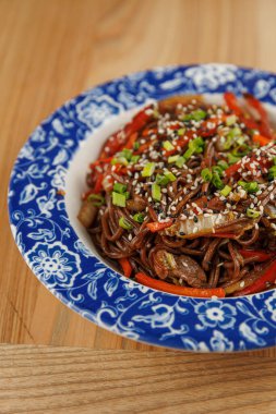 Soba erişteleri üzerine susam tohumu ve yeşil soğan konmuş mavi ve beyaz bir kâse ile kaplıdır. Parlak sebzeler bu yemeğe sıcak mutfak ortamında renk katar..