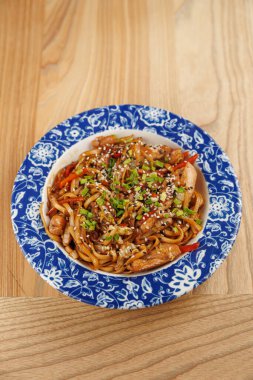 Farklı mutfaklardan gelen çeşitli yemek tabakları ahşap bir masanın üzerindeki renkli bir kaseye konur. Yemekler erişte, sebze, tohum ve düzgün bir şekilde servis edilen etleri içeriyor..