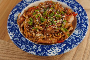 Bir kasede tavuk, sebze ve yeşil soğan ve susam tohumlarıyla karıştırılmış kızarmış erişte var. Sahne, kültürel tatların bir karışımını yakalıyor..