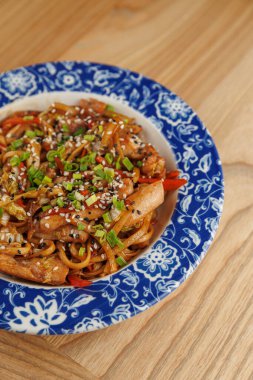 Üzerine tavuk parçaları ve renkli sebzeler ile karıştırılmış erişte ahşap bir masada mavi desenli bir kasede servis edilir. Yemeklerde susam tohumları ve süslemeler var..