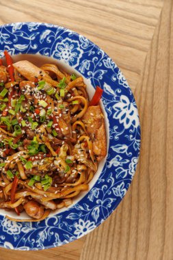 Tavuklu ve sebzeli bir kase kızarmış şehriye mavi dekoratif bir tabakta duruyor. Yemek susam tohumları ve doğranmış yeşil soğanla süslenmiş, yemeye hazır..