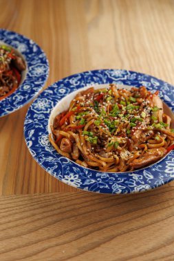 Renkli erişte, sebze ve tavukla dolu iki kase ahşap bir masada oturur. Dilimlenmiş otlar ve susam tohumları tabağın üstünde. Aydınlık ve davetkar bir ortam..