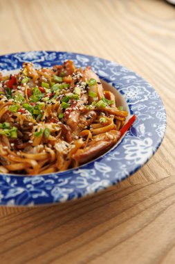 Bir kase kızarmış şehriye renkli sebzelerle doldurulur ve susam tohumları ve doğranmış yeşil soğanla süslenir. Tabak ahşap bir masaya kurulmuş, canlı sunumunu vurguluyor..