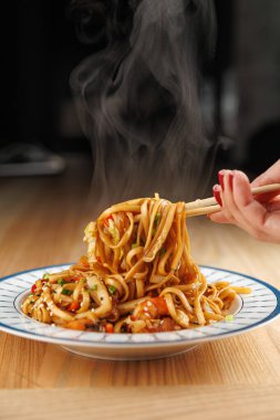 A hand holds chopsticks picking up steaming noodles from a plate. The dish includes thick noodles mixed with vegetables. A warm kitchen ambiance enhances the scene.