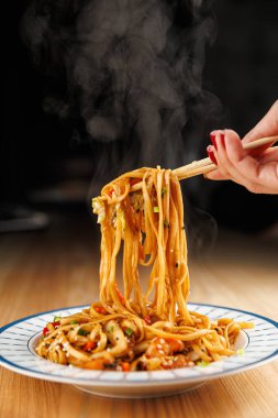 A person holds chopsticks picking up steaming noodles from a plate filled with colorful vegetables. The scene captures the enjoyment of a flavorful dish in a cozy atmosphere.
