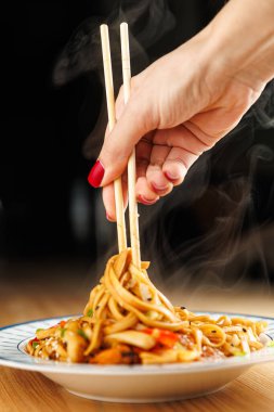 A hand with red nails uses chopsticks to lift steaming noodles from a plate. The dish features vibrant vegetables and fragrant sauce, showcasing a diverse meal from Asian cuisine.