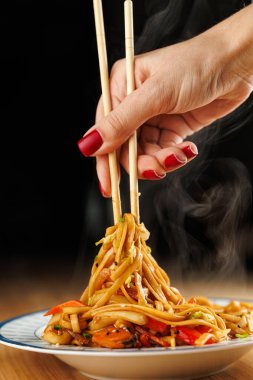 A hand holds chopsticks, picking up a mound of steaming noodles mixed with vegetables. The scene highlights the enjoyment of Asian cuisine in a cozy atmosphere.