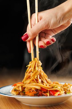 A hand holding chopsticks lifts steaming noodles from a plate filled with vibrant vegetables. The close-up captures the movement of food and the warmth of the dish.