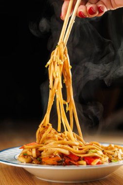 A hand holds chopsticks lifting steaming noodles from a plate filled with mixed vegetables. The warm dish is perfect for sharing.