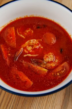 Deniz ürünleri, domatesler ve diğer sebzelerle dolu bir kase kırmızı çorba ahşap bir masaya konur. Çorba lezzetli ve lezzetli görünüyor, her yemeğin tadını çıkarmaya hazır..