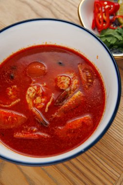 Bir kase zengin kırmızı deniz ürünleri çorbası ahşap bir masanın üzerine konur. Tabakta karides ve baharatlı et suyuyla çevrili diğer deniz ürünleri yer alıyor. Kırmızı biberli ve otlu bir tabak yemeği tamamlar..