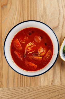 Karides ve sebzeli zengin kırmızı bir çorba ahşap bir masada bir kaseye konur. Çorbada çeşitli tatlar ve parlak renkler var..