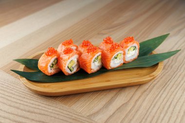 Somon, avokado ve pirinçle dolu suşi ruloları ahşap bir tabakta düzenlenir. Ekmekler turuncu balık yumurtası ile kaplı ve yeşil bir yaprak üzerinde sunuluyor..