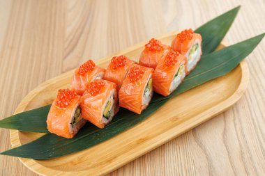 Somon ve yumurtayla yapılan suşi ruloları yeşil yapraklı ahşap bir tahtaya yerleştirilir. Yemek ustaca hazırlanmış suşi hazırlama ve sunumu vurguluyor.