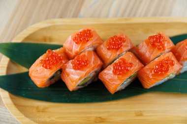 Taze hazırlanmış suşi ruloları ahşap bir tabakta sunulur. Her rulo somon dilimleri içeriyor ve üzerinde parlak kırmızı balık yumurtası var. Yeşil yapraklar renk ekler.