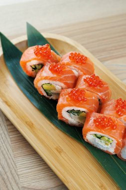 Parlak suşi ruloları ahşap bir tabakta güzelce düzenlenir. Bu rulolarda somon, avokado ve süslemeler yer alıyor. Modern bir ortamda yemeğin canlı renklerini vurguluyorlar..