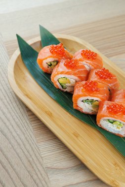 Suşi ruloları bambu bir tabakta güzelce sıralanır, üzerinde somon dilimleri ve renkli dolgular bulunur. Tabak dekoratif bir yaprak üzerinde servis edilir..