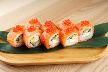 Suşi ruloları bambu tepsisinin üzerine güzelce dizilir. Her rulo somon, avokado ve parlak yumurtayla kaplı salatalık içeriyor. Yeşil yapraklar sunum için temel olarak kullanılır.