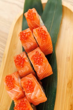 Parlak turuncu yumurtayla kaplanmış somon balığı parçaları ahşap bir tabağa yerleştirilmiş yeşil bir yaprağa yerleştirilmiş ve bir yemek masasında canlı bir yemek sunumu sergilenmektedir..